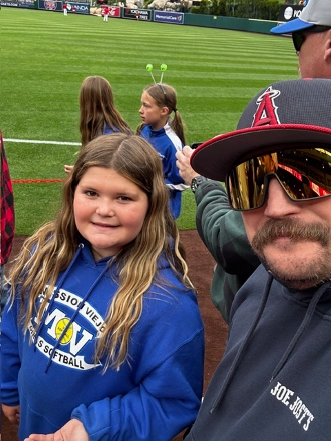 A girl in a blue sweatshirt smiles next to a man wearing sunglasses at a baseball game.