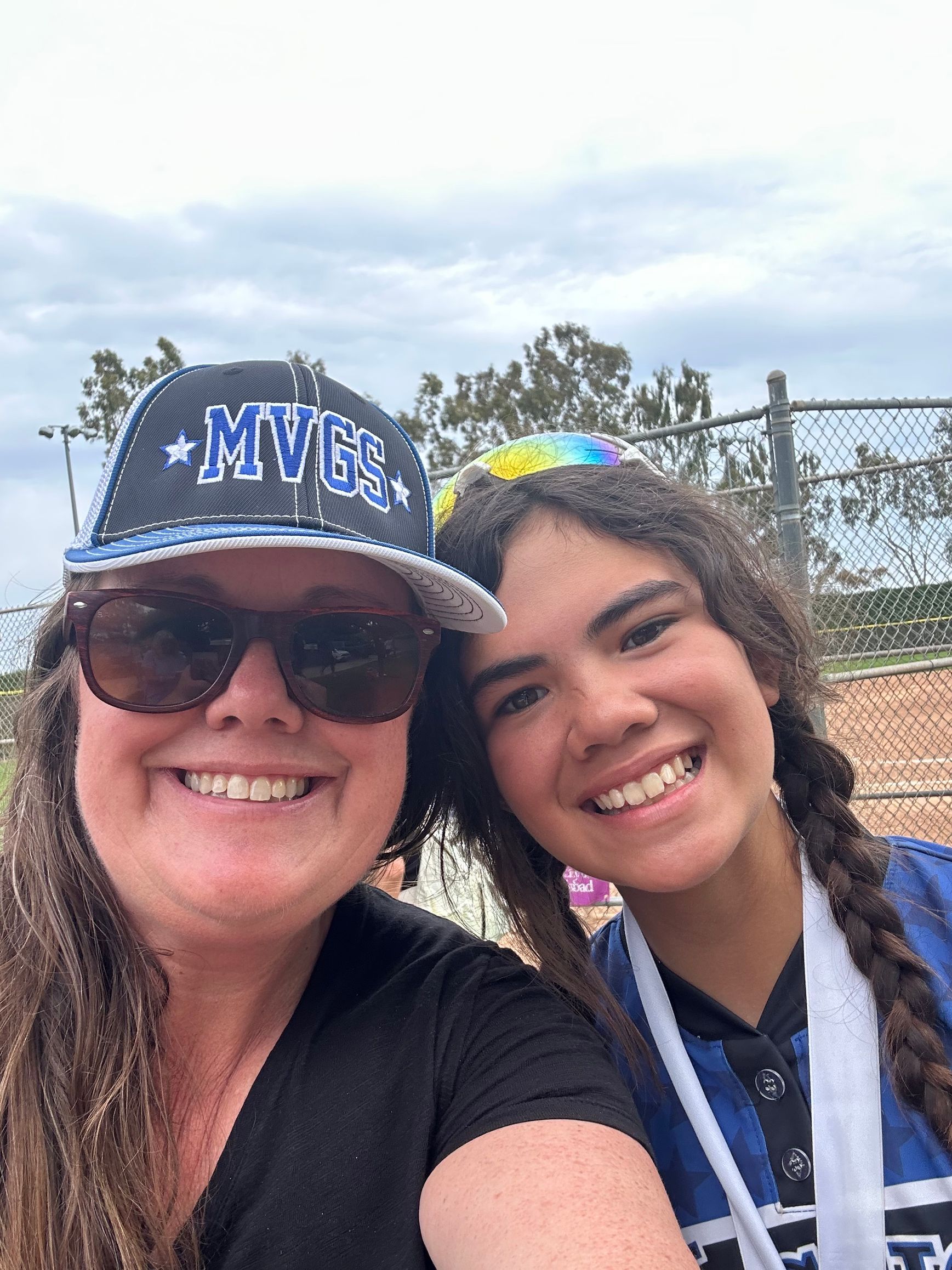 Two smiling people: One wearing sunglasses and a baseball cap, the other with a braid and a medal, posing outdoors.