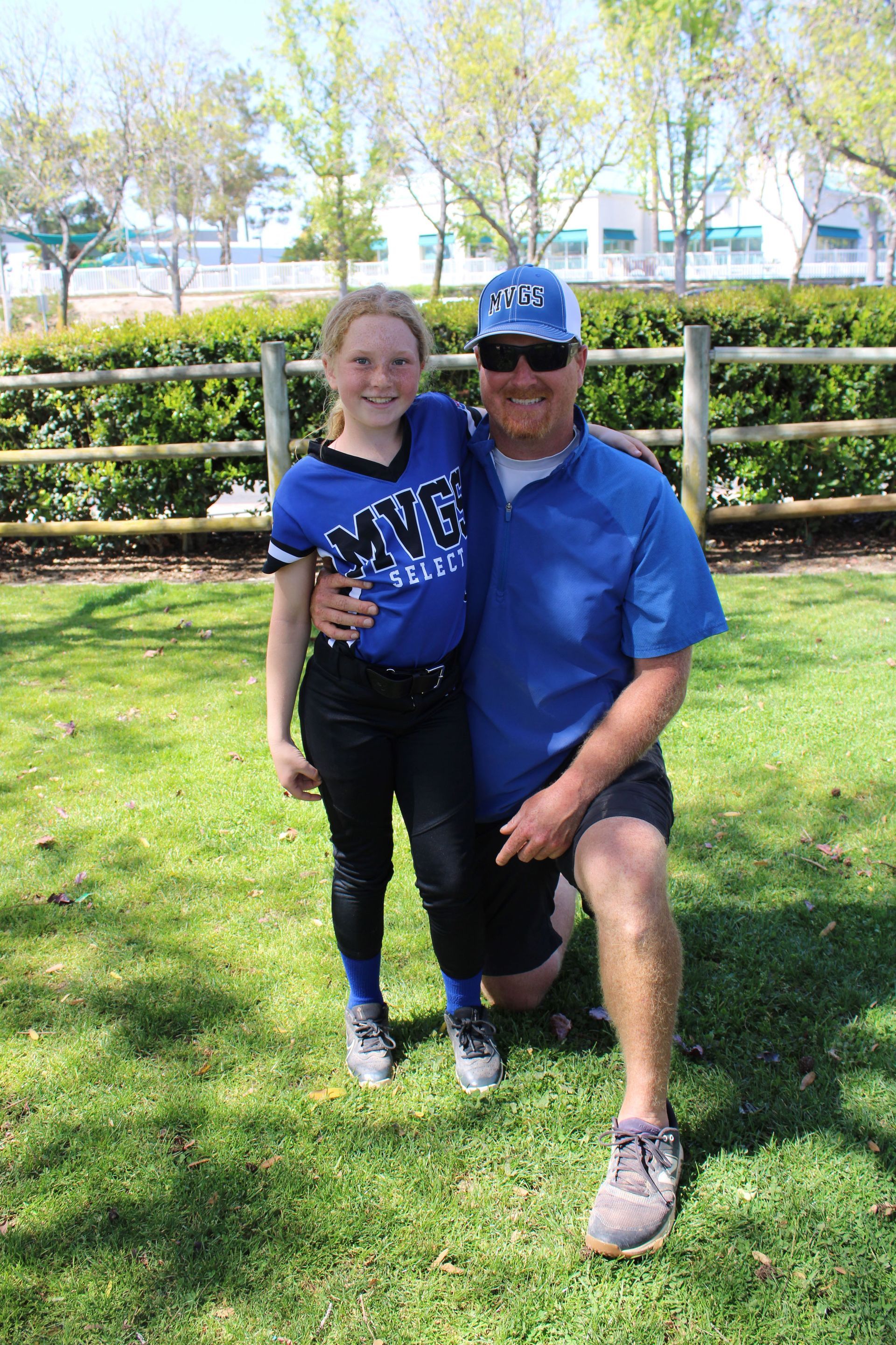 A girl in a blue softball uniform and a man in a blue shirt pose outside. The man kneels on the grass.