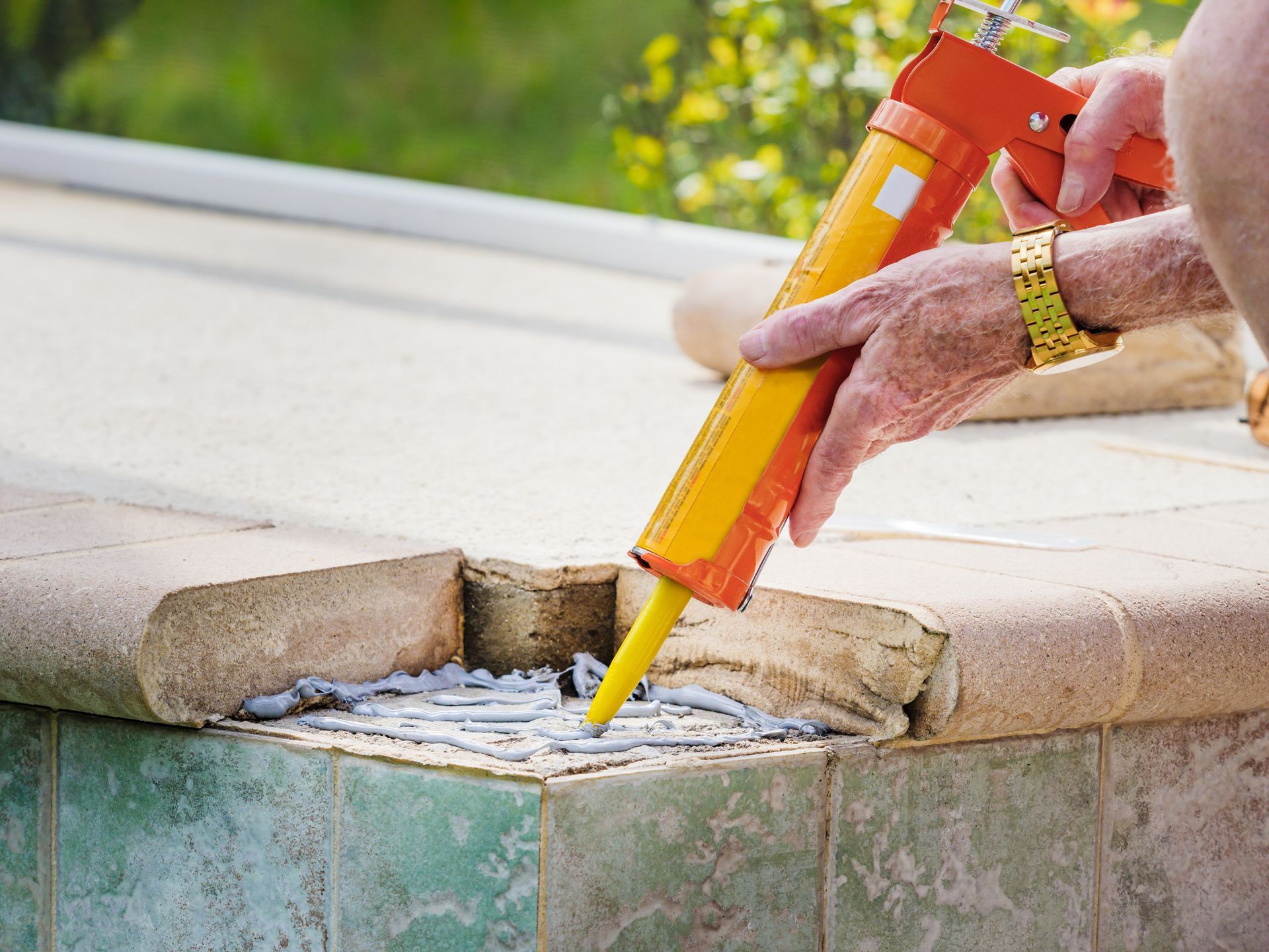 Person applying caulk to a damaged pool edge with an orange and yellow caulk gun.