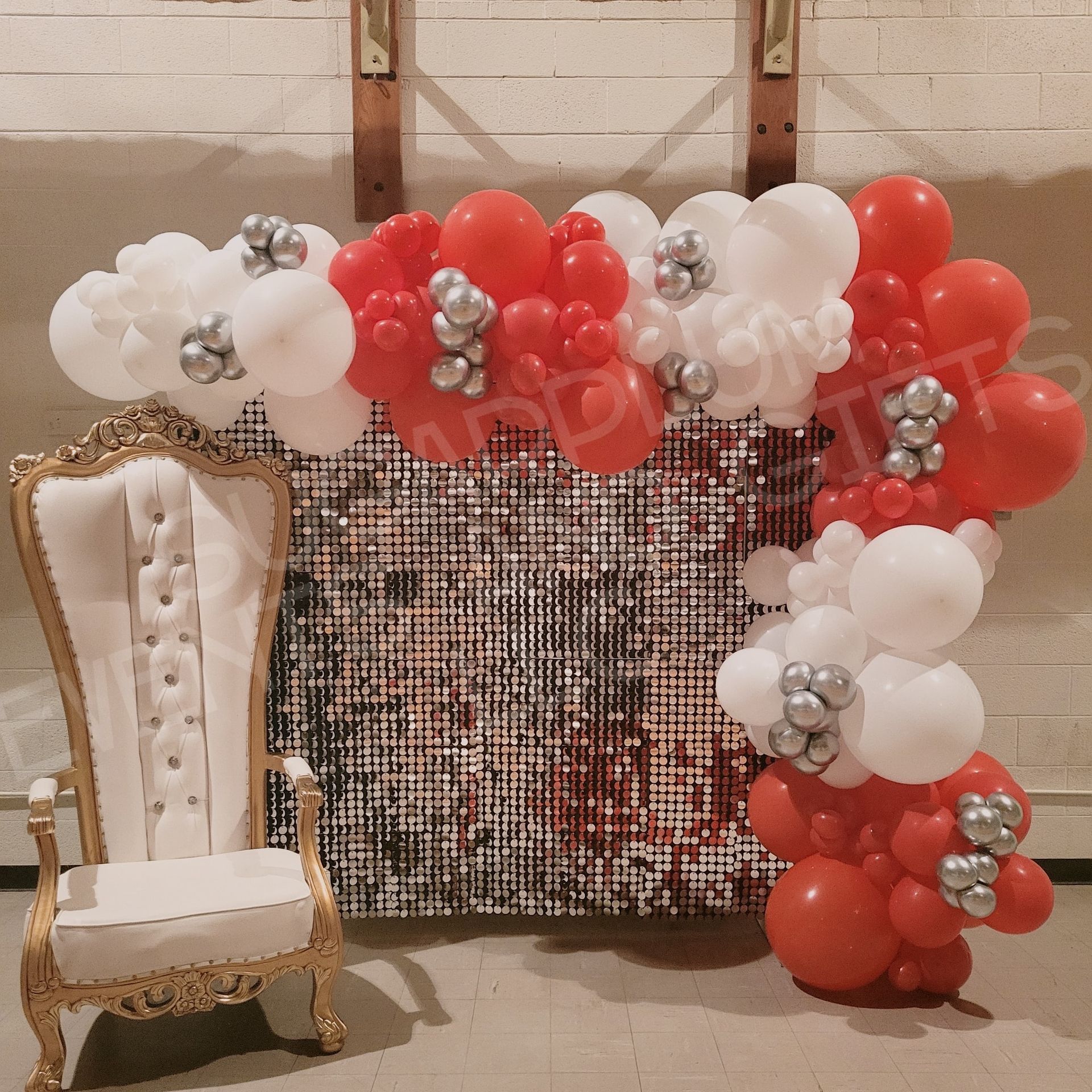 Elegant white chair beside red, white, and silver balloon arch in front of a sequined backdrop