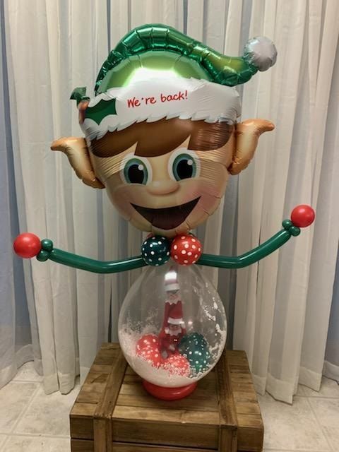 Elf holiday decoration holding an hourglass, wearing a green hat and red gloves, standing on a wooden crate