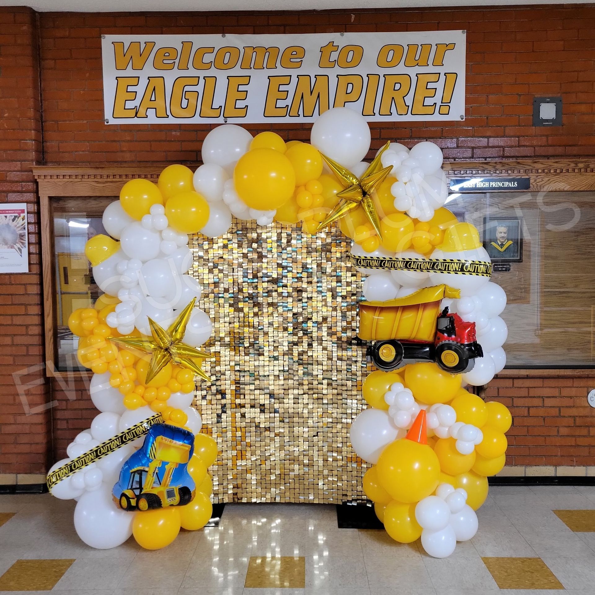 School hallway display with gold and white balloons, “Welcome to our EAGLE EMPIRE!” sign, and toy trucks on a gold backdrop
