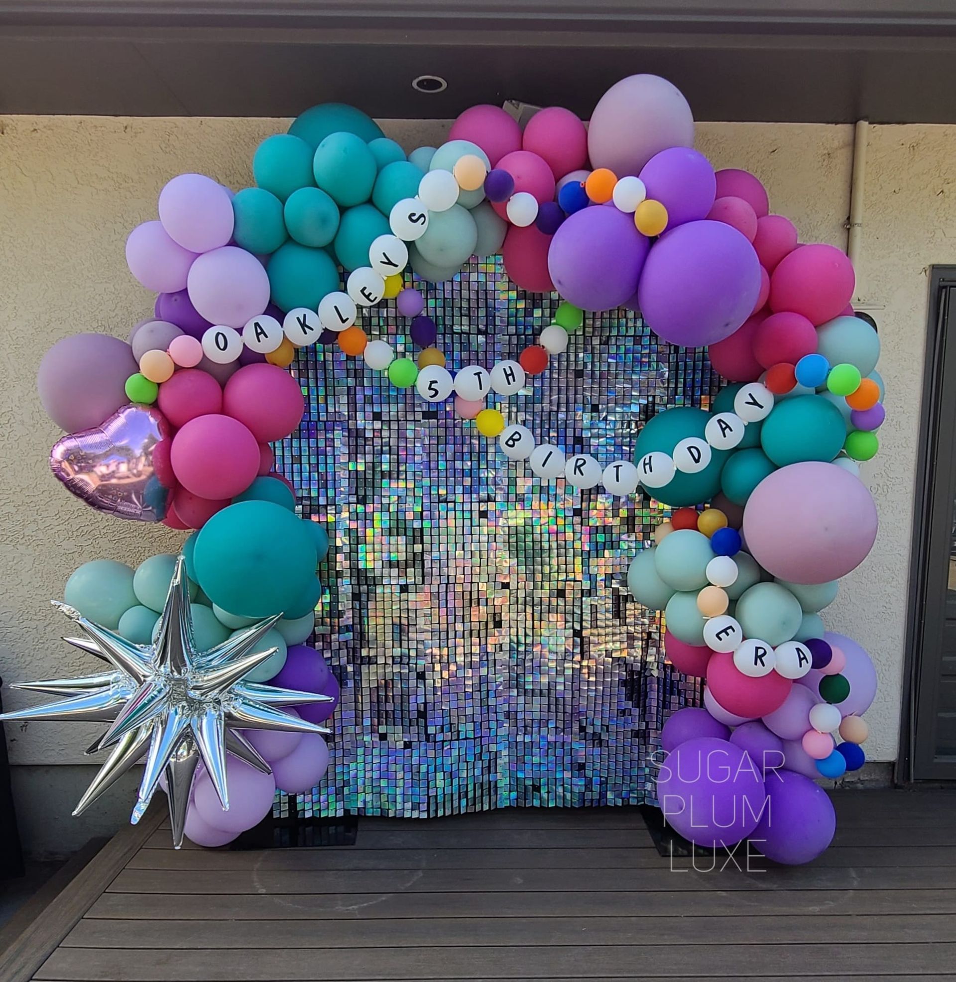 Pastel balloon arch framing a shimmering sequin backdrop with star props and purple name balloons on a wall