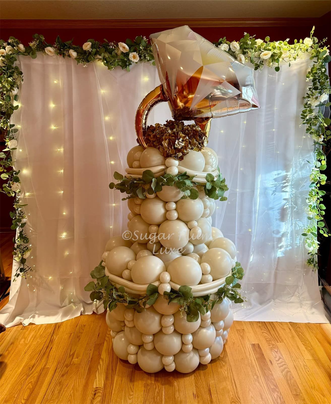 Balloon tower centerpiece with white and gold balloons, greenery, and a gift bag on a draped backdrop