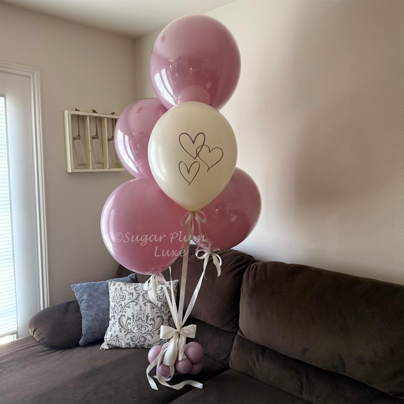Pink and cream heart balloons with a gift bag beside a dark sectional sofa in a living room