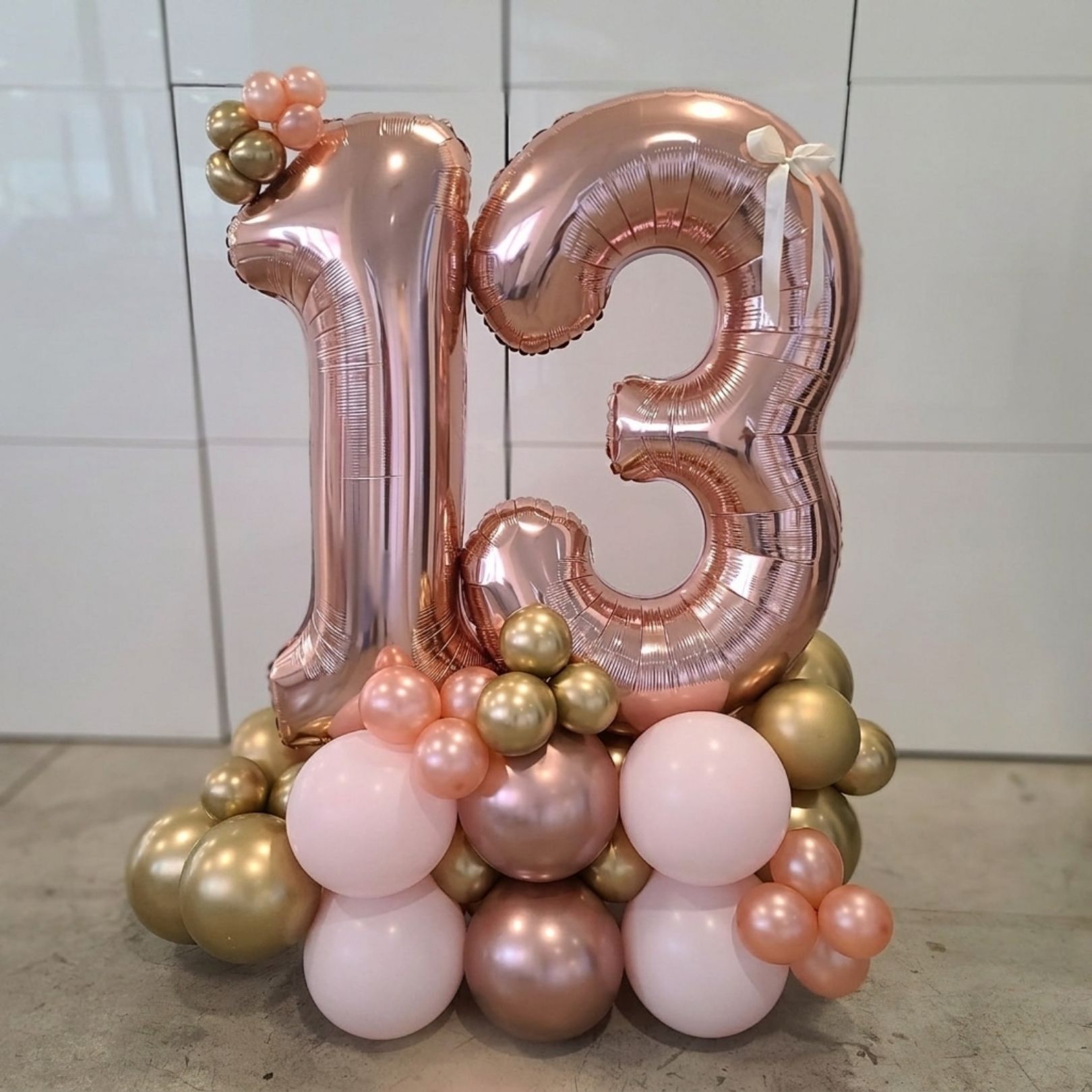 Rose gold and pink balloon display with a large number 13 and floral accents on a tiled floor