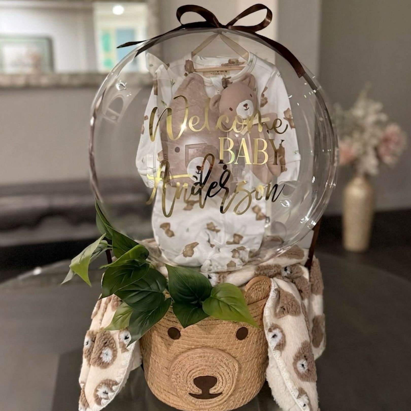 Clear balloon baby shower centerpiece with gold lettering, teddy bear base, and greenery on a table.