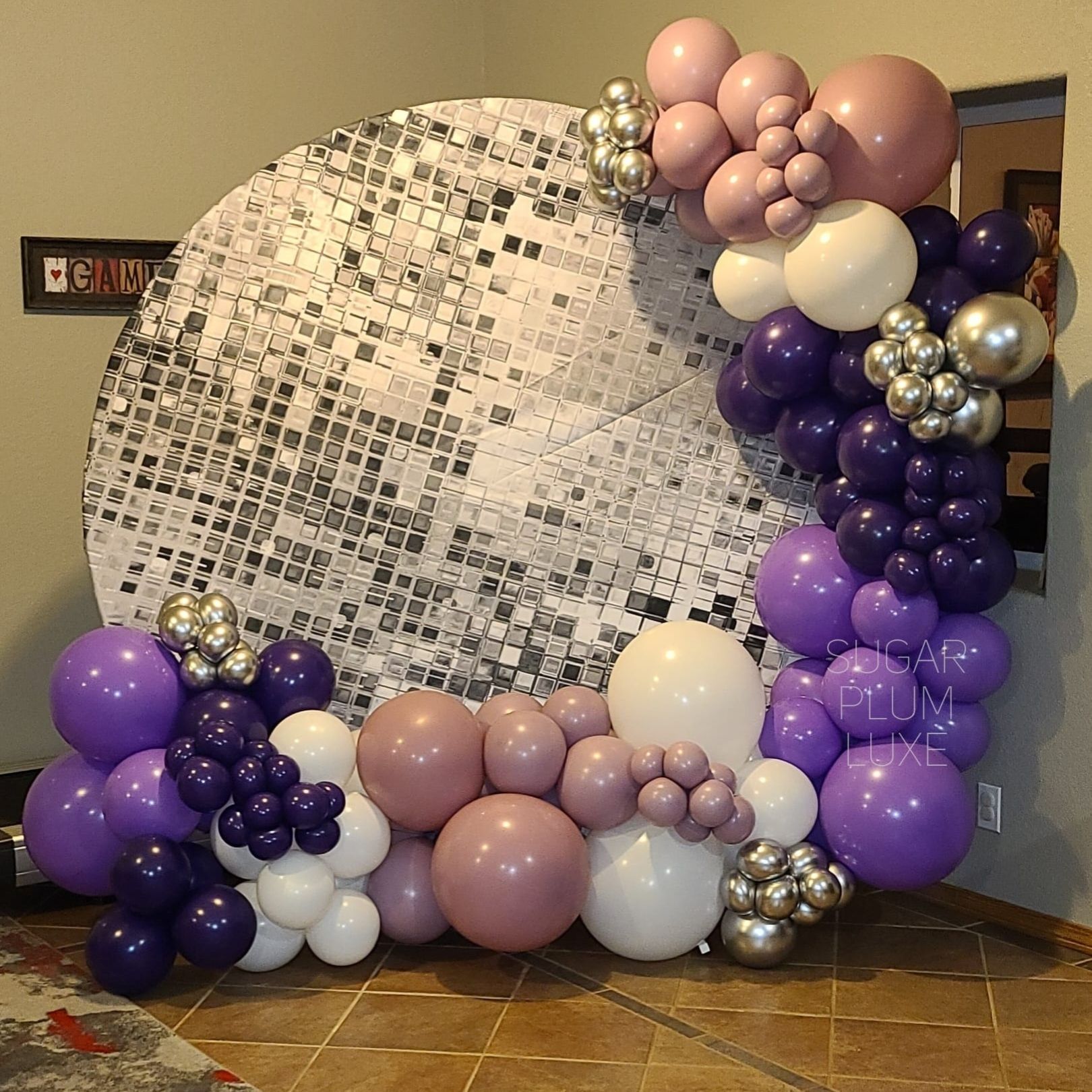 Purple, silver, and blush balloon arch framing a large circular mosaic wall at an indoor event.