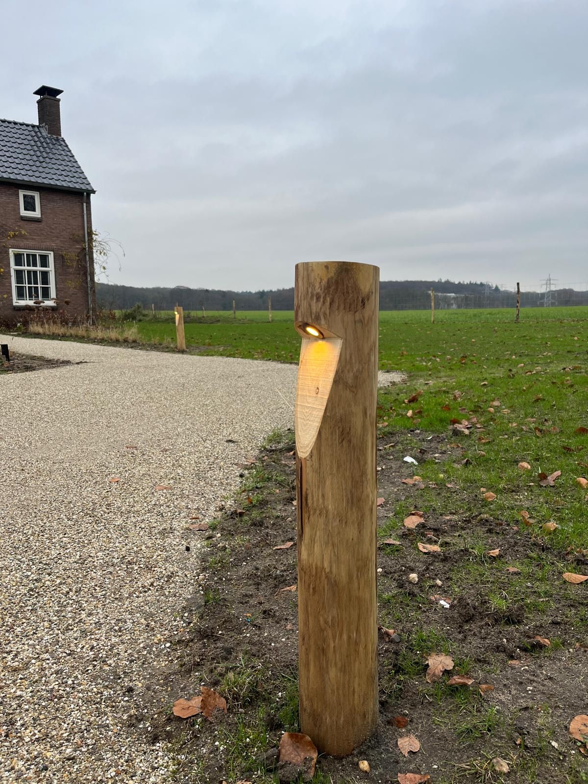 Houten paal met ingebouwde lamp, naast een grindpad en veld.