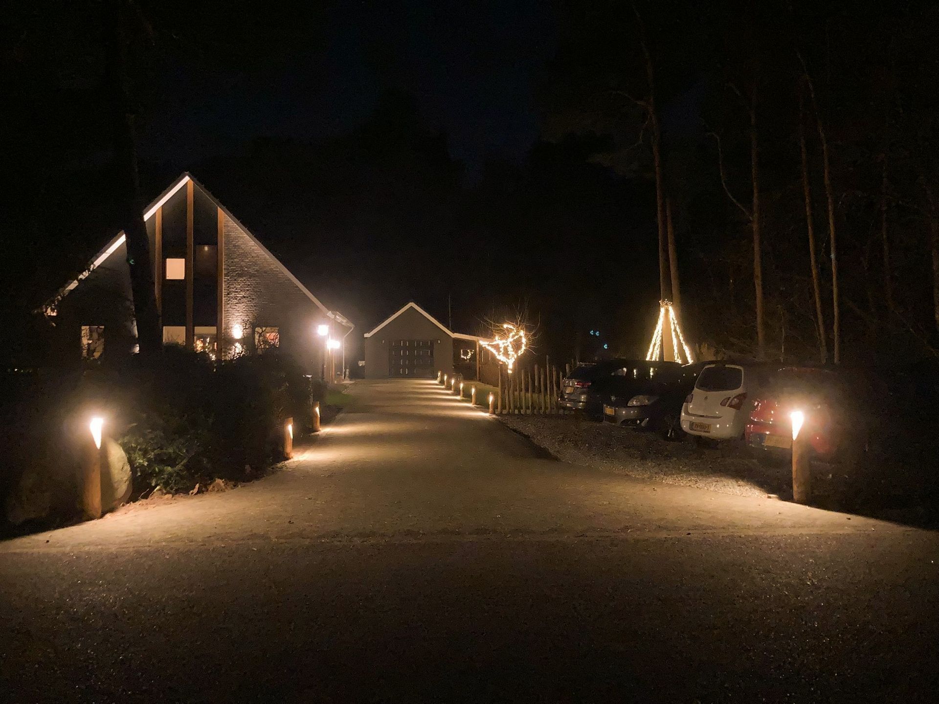 Nachtscène van een gebouw en een pad met verlichting, feestelijke versieringen en geparkeerde auto's, onder een donkere hemel.