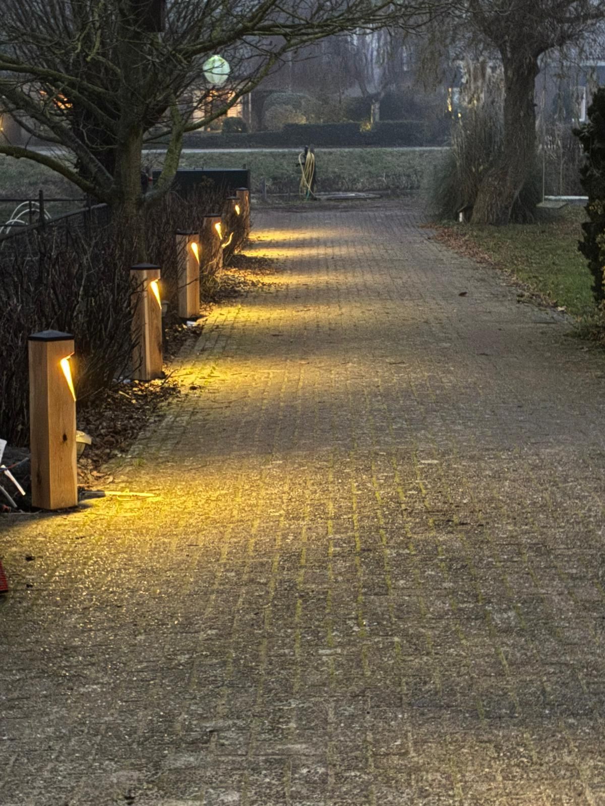 Een pad in een parkachtige omgeving wordt verlicht door een rij lampen. Het warme licht weerkaatst op de bestrating.