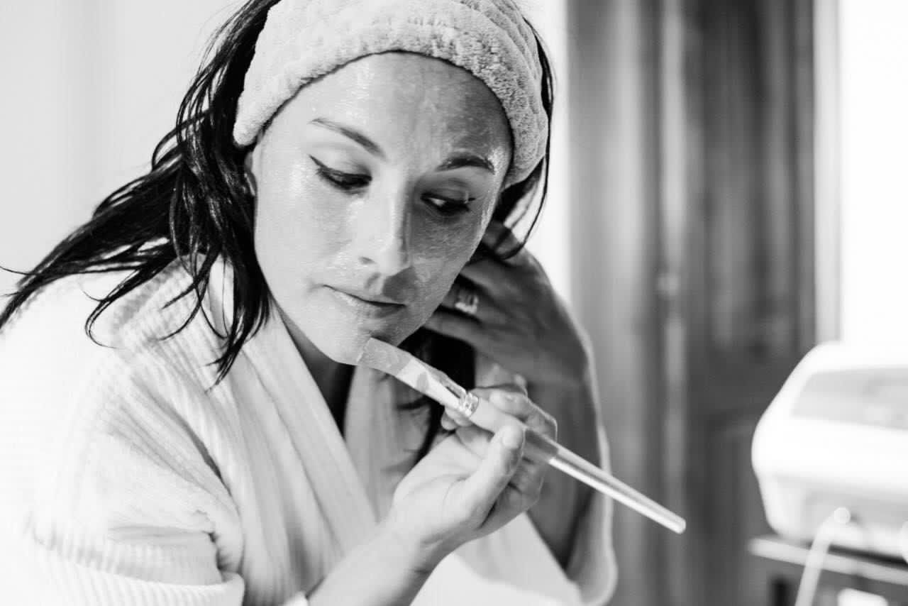 Mujer en bata de baño, aplicándose una mascarilla facial con un palo de madera, diadema, entorno de spa.