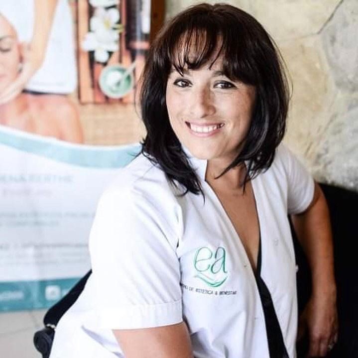 Mujer con bata blanca sonríe, probablemente en un spa, con un cartel y una bolsa negra visibles.