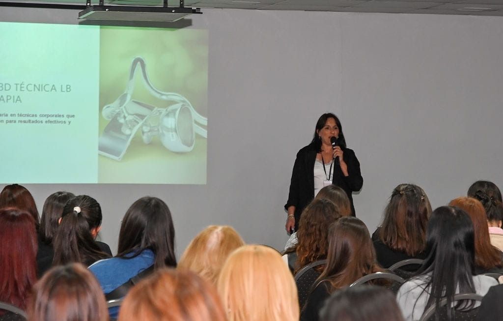 Mujer dando una presentación a una audiencia sentada, con una pantalla de proyector en el fondo.