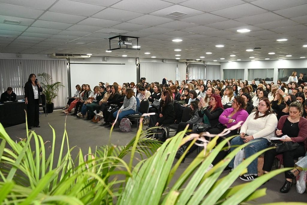 Mujer hablando ante una gran audiencia en una sala de conferencias; plantas verdes en primer plano.