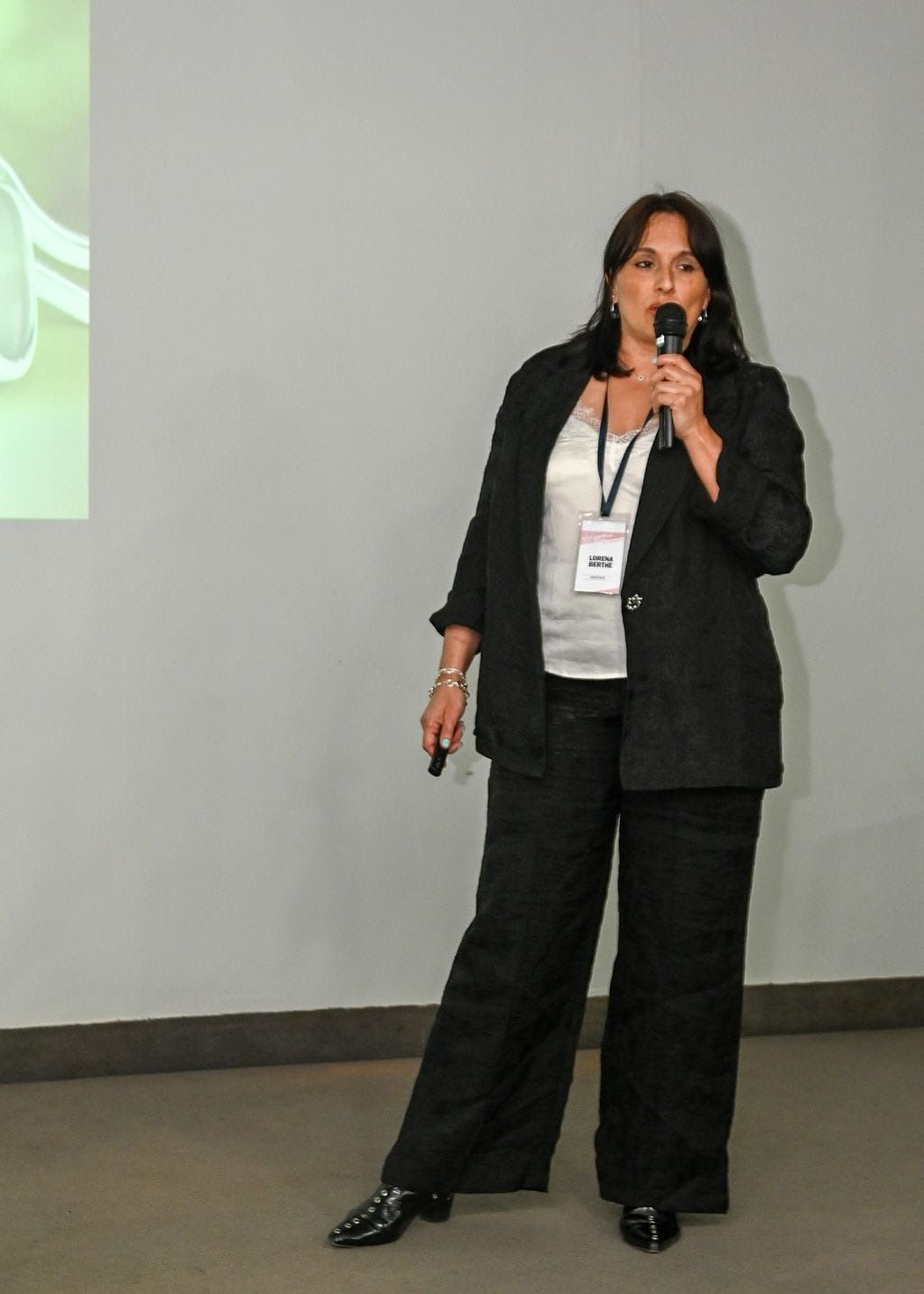 Una mujer con traje negro habla por un micrófono mientras hace una presentación sobre un fondo de pared liso.