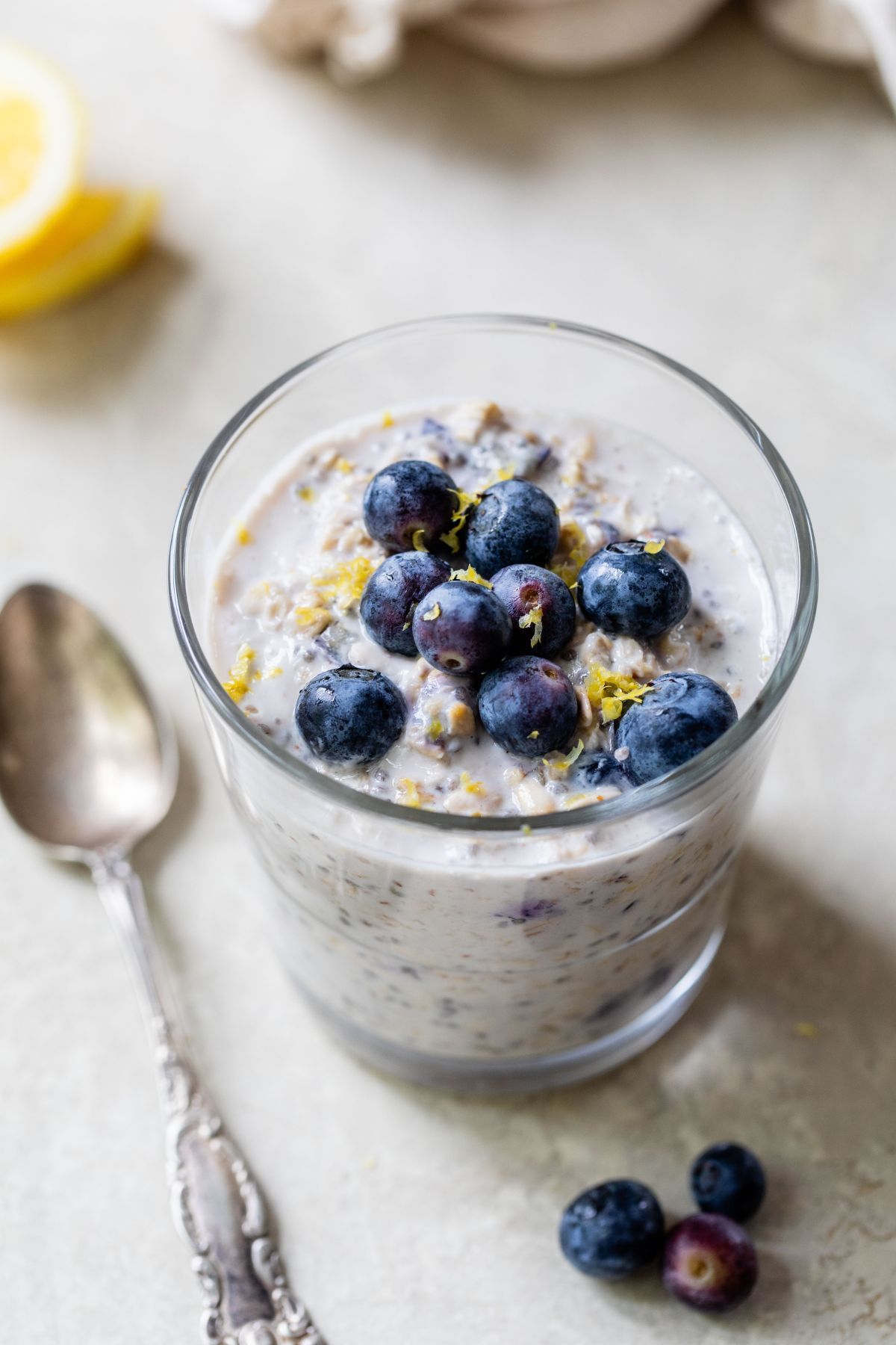Glass of blueberry oatmeal topped with zest and fresh blueberries, beside a spoon.