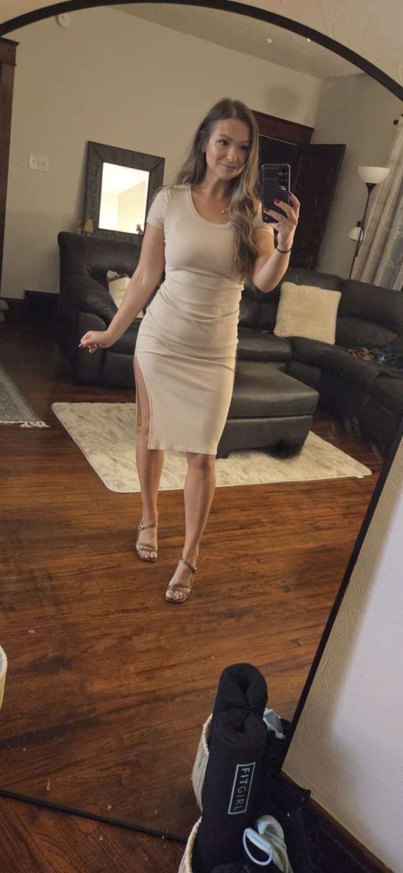 Woman in a light beige dress poses in a mirror, holding a phone. She is in a living room with wooden floors.