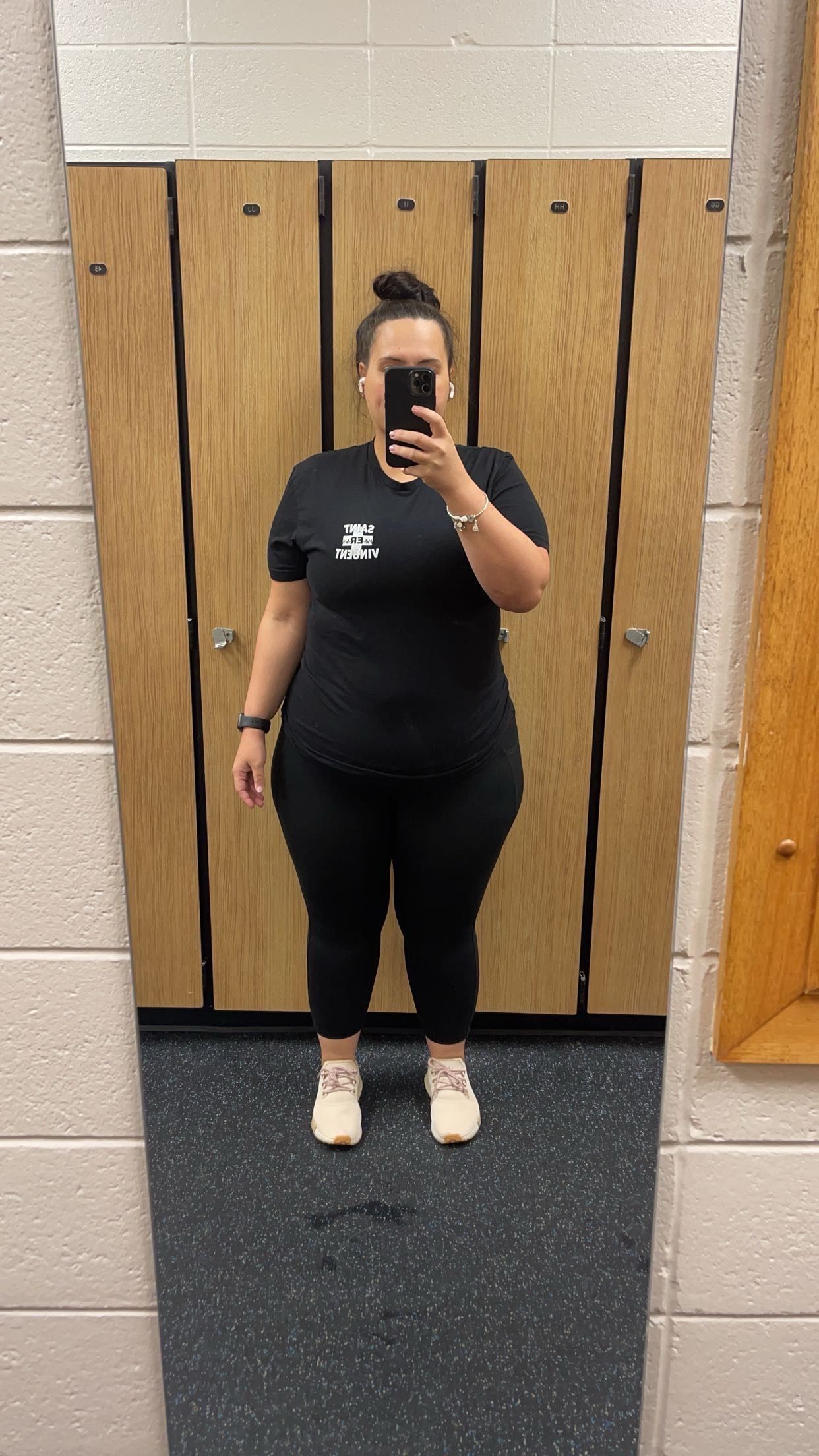 Person in black athletic wear stands in front of a mirror, holding a phone, inside a locker room.