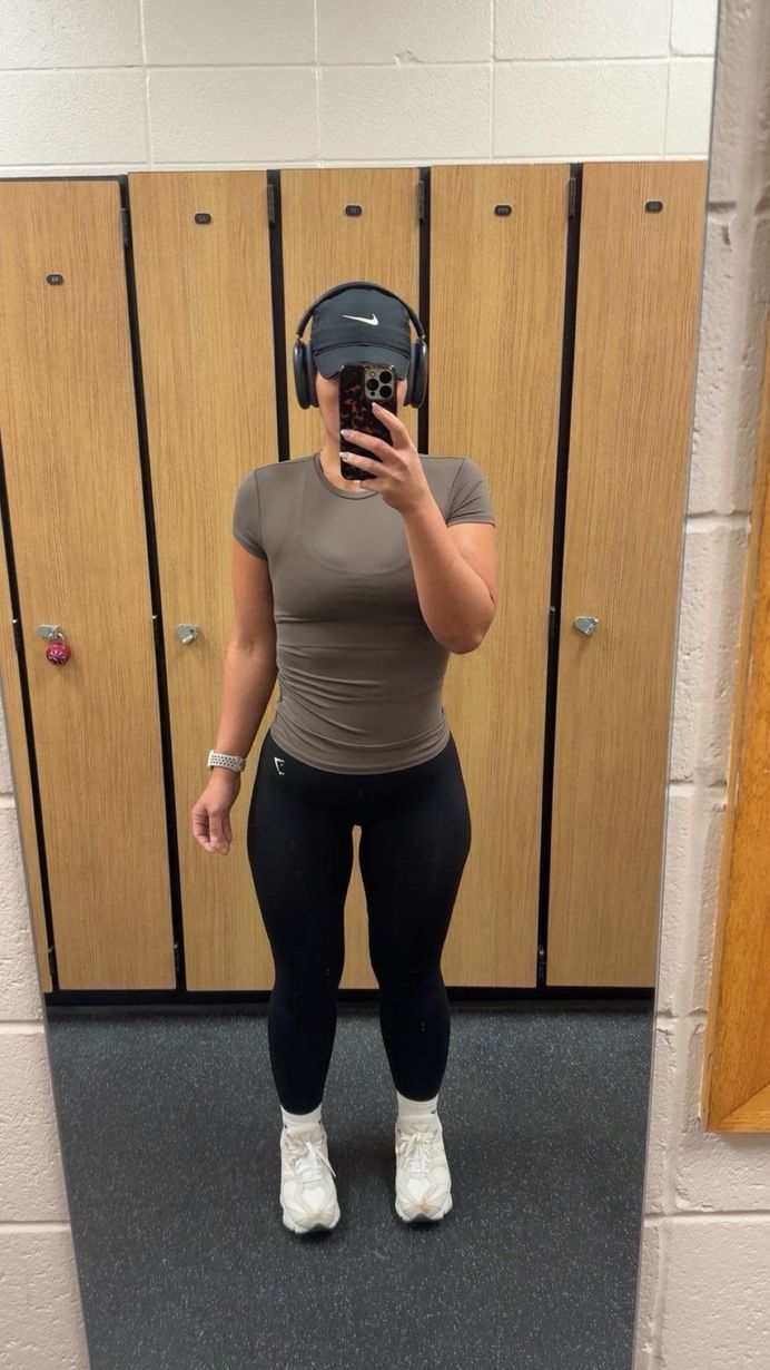 Woman in gym attire takes a mirror selfie.  She wears a gray shirt, black leggings, and white sneakers, with headphones.