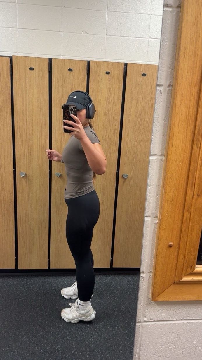 Woman in gym clothes in front of a mirror in a locker room, holding phone.