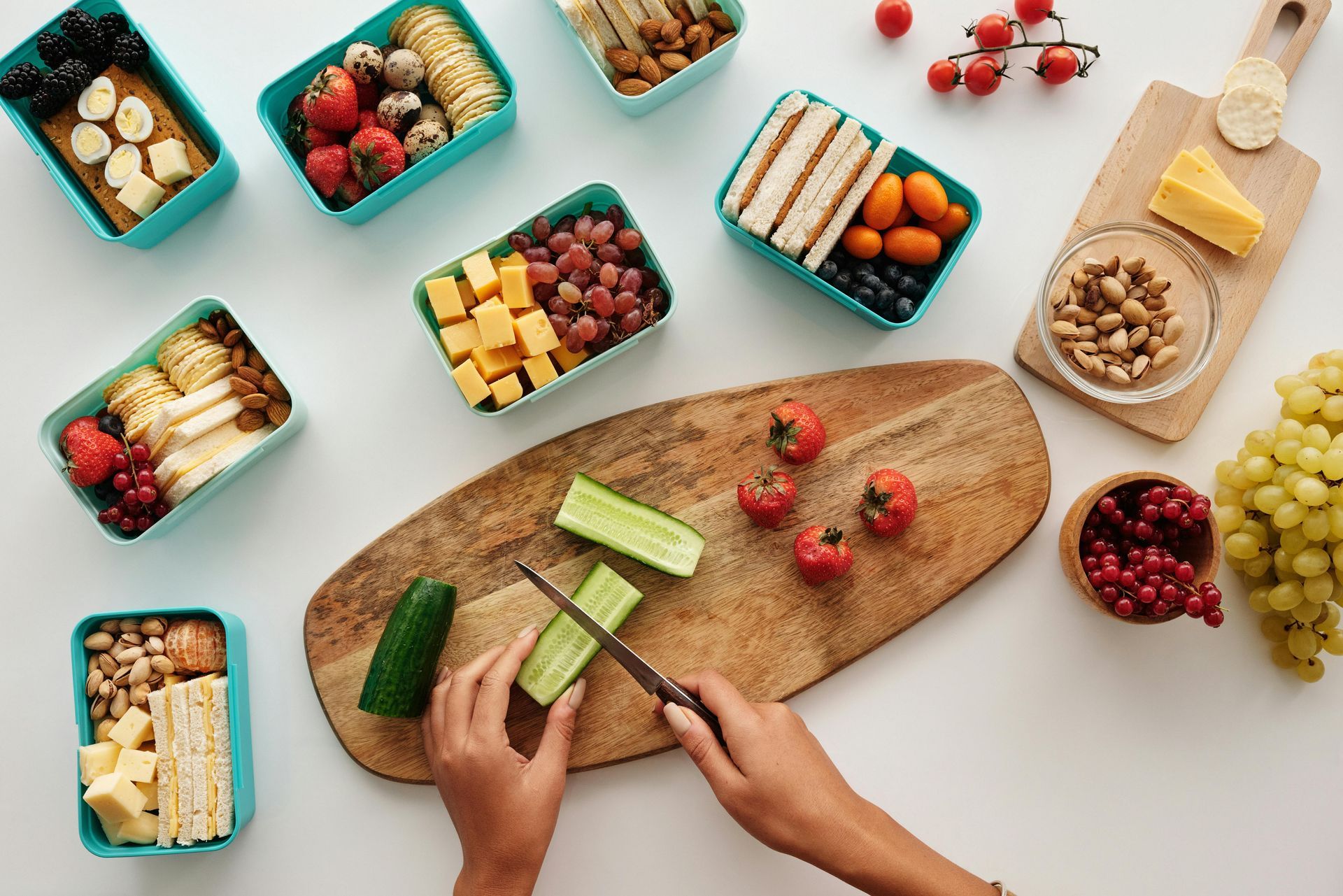 Person cutting cucumber on a wooden board surrounded by packed lunches with fruits, cheese, and crackers.
