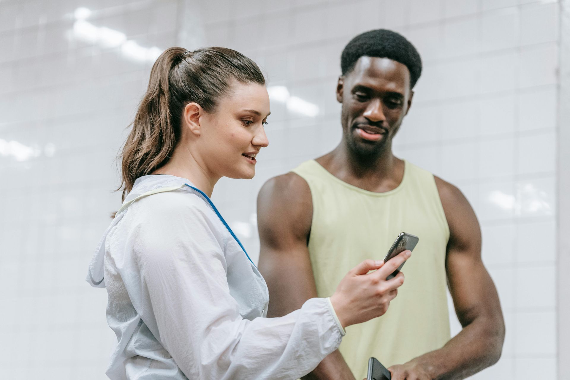Woman in white jacket and man in yellow tank top looking at a phone in a gym setting.