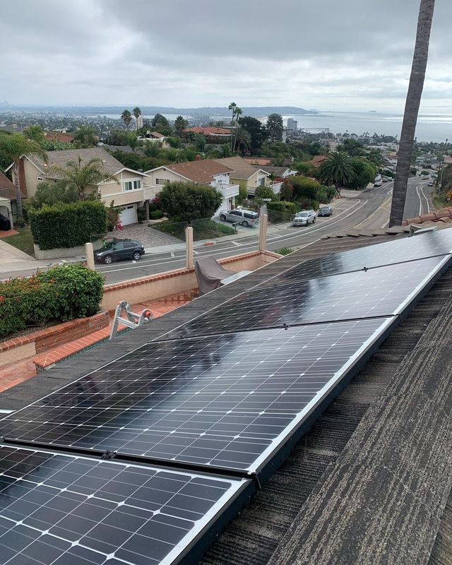Solar panels on a rooftop overlooking a suburban neighborhood and distant water under a cloudy sky