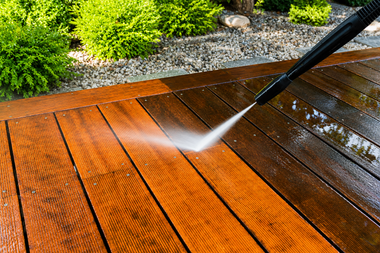 Pressure washing a wooden deck beside green shrubs, spraying off dirt and water.