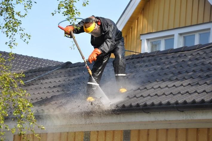 Worker using a power tool on a house roof, creating dust near a yellow exterior wall.