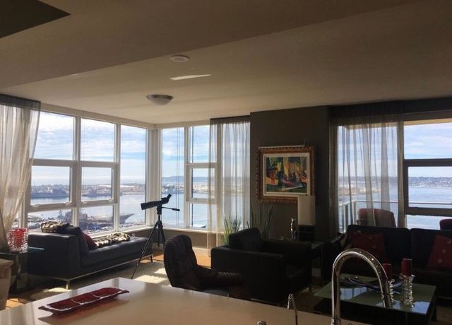 Sunlit living room with floor-to-ceiling windows overlooking water, dark sofas, and a telescope