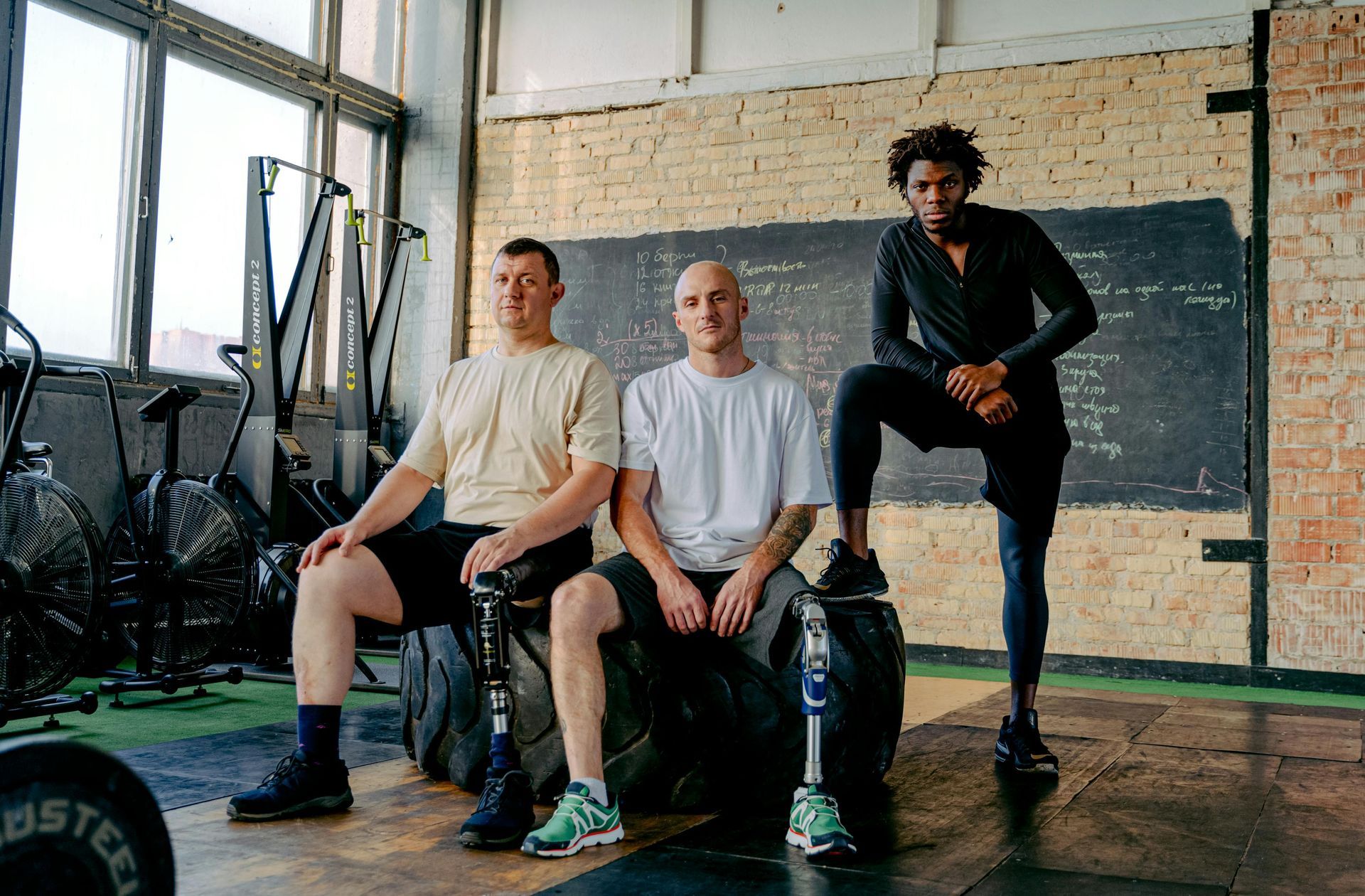 Three men at a gym. Two seated on tires, each with a prosthetic leg. The other stands, leaning on a tire, smiling.