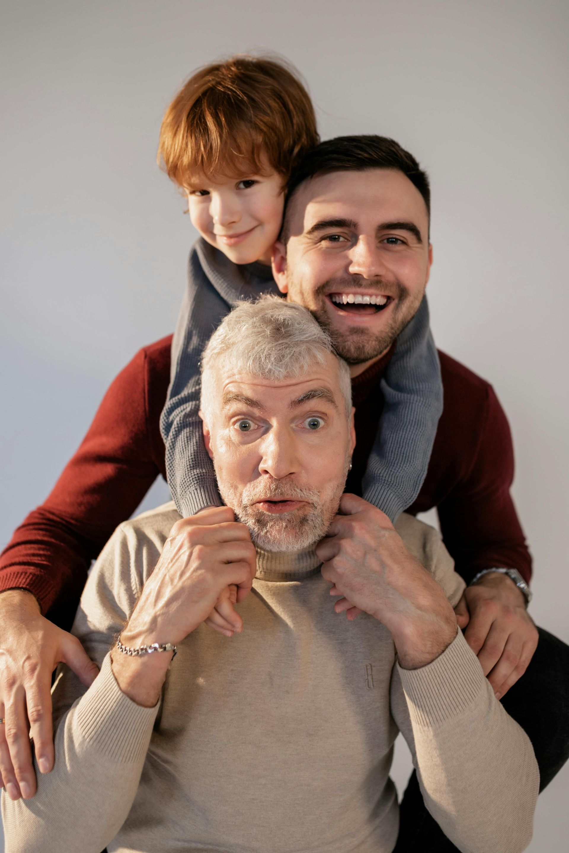 Three generations, a boy on his father's shoulders, dad on grandpa's. All smiling. Against a white background.