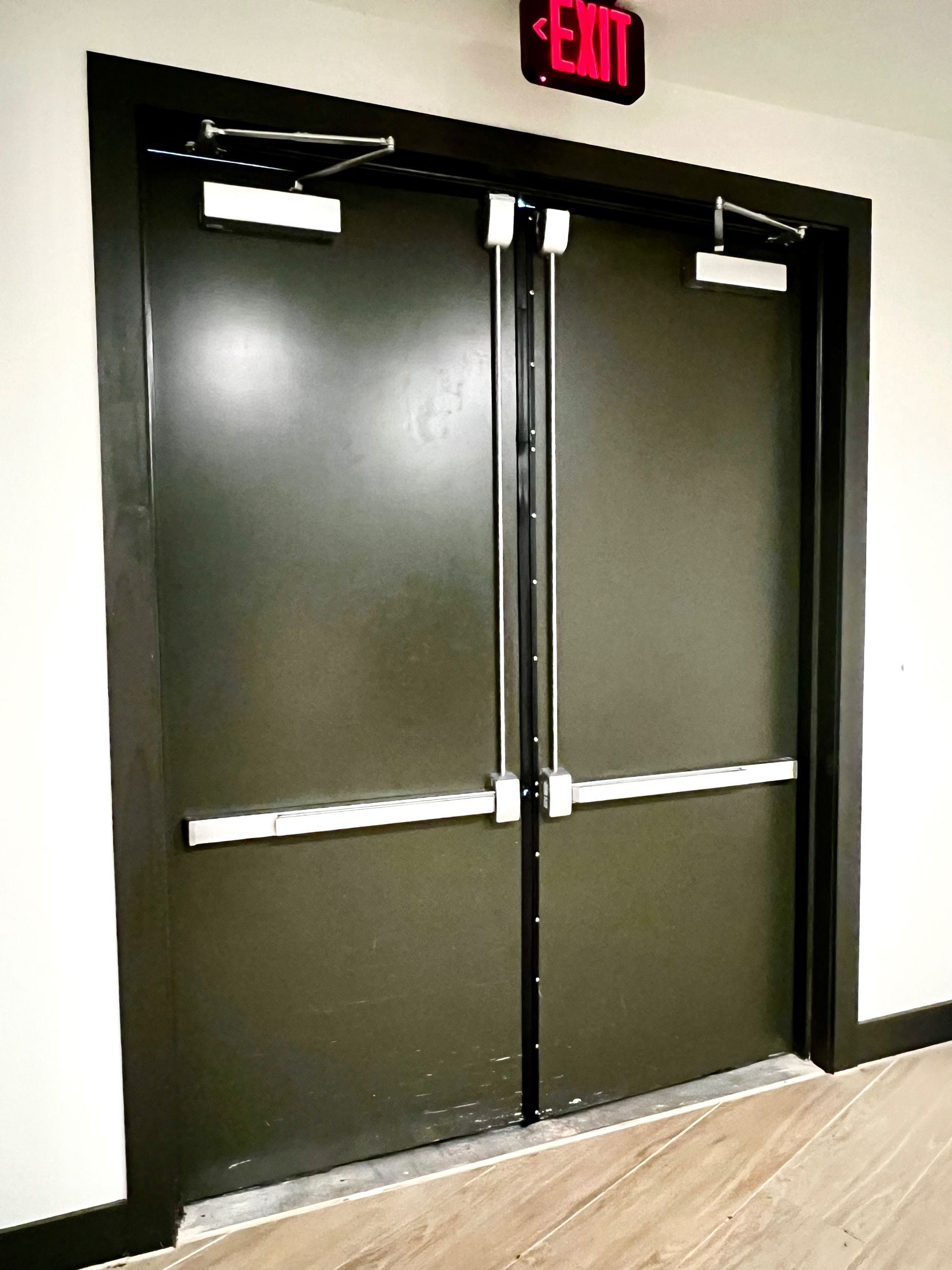 Black double exit doors with push bars and red EXIT sign in a hallway.
