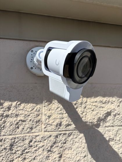 White security camera mounted on a beige exterior wall, casting a long shadow.