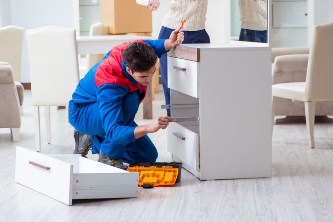 Person in blue work clothes assembling a white cabinet in a bright living room, with tools on the floor