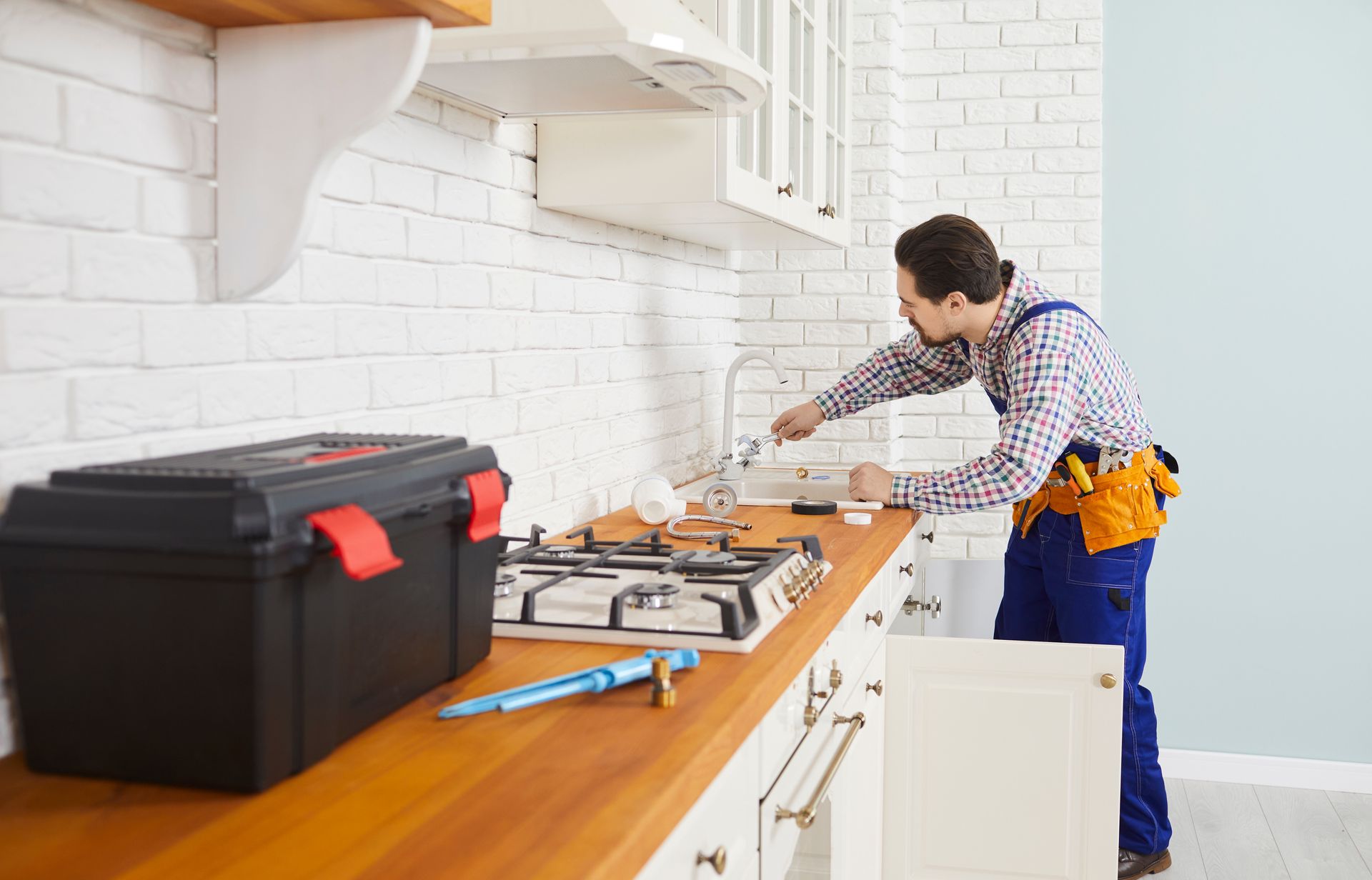 Man fixing a kitchen faucet in a white kitchen, with tools on a wooden counter and a black toolbox nearby