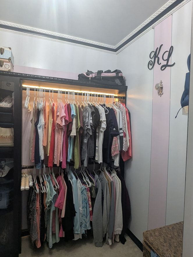 Closet with rows of hanging clothes, lit shelf, and mirror in a small room