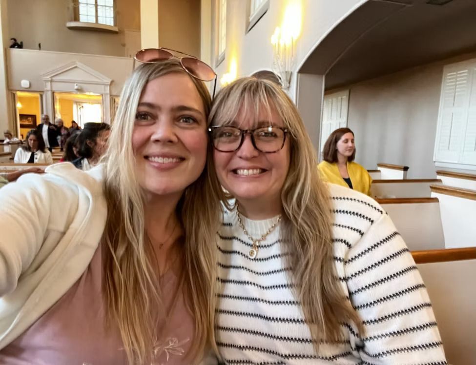 Two women smiling in a light-filled room with pews, one wearing sunglasses on her head.