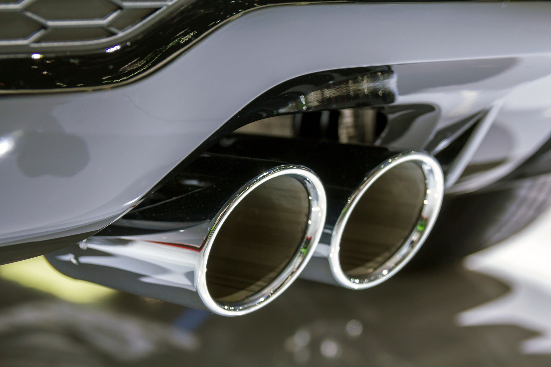 Close-up of a car’s chrome dual exhaust pipes beneath a gray rear bumper Close-up of a car’s chrome dual exhaust pipes beneath a gray rear bumper