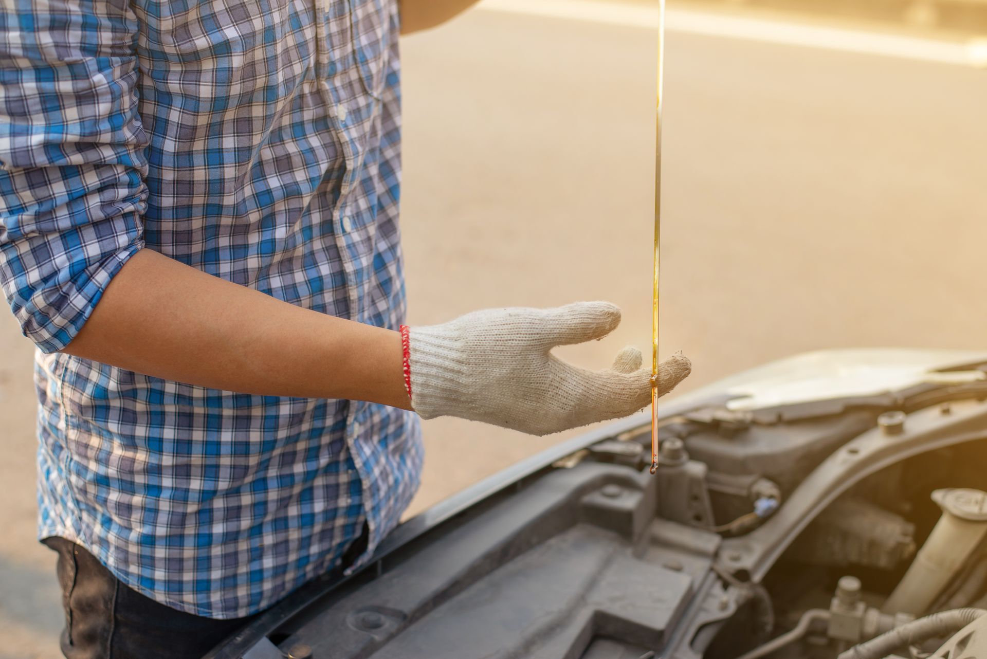 Person in a blue plaid shirt checking a car’s oil with a dipstick under the open hood