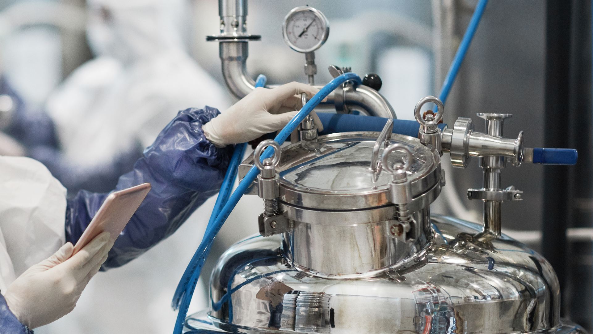 Gloved technician adjusts tubing on a stainless steel lab reactor with gauges and blue hoses