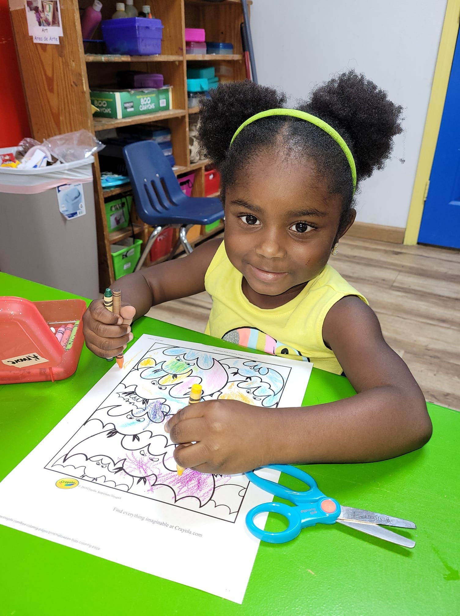 A little girl is sitting at a table with scissors and a pencil.