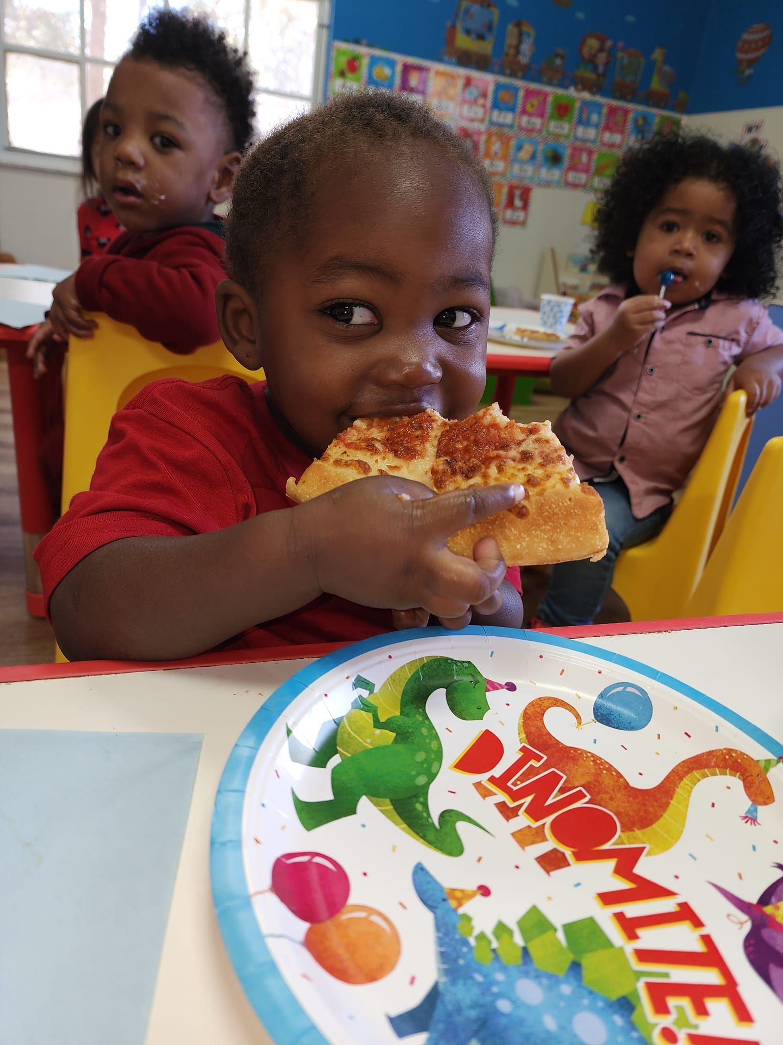 A little boy is eating a slice of pizza next to a paper plate with dinosaurs on it