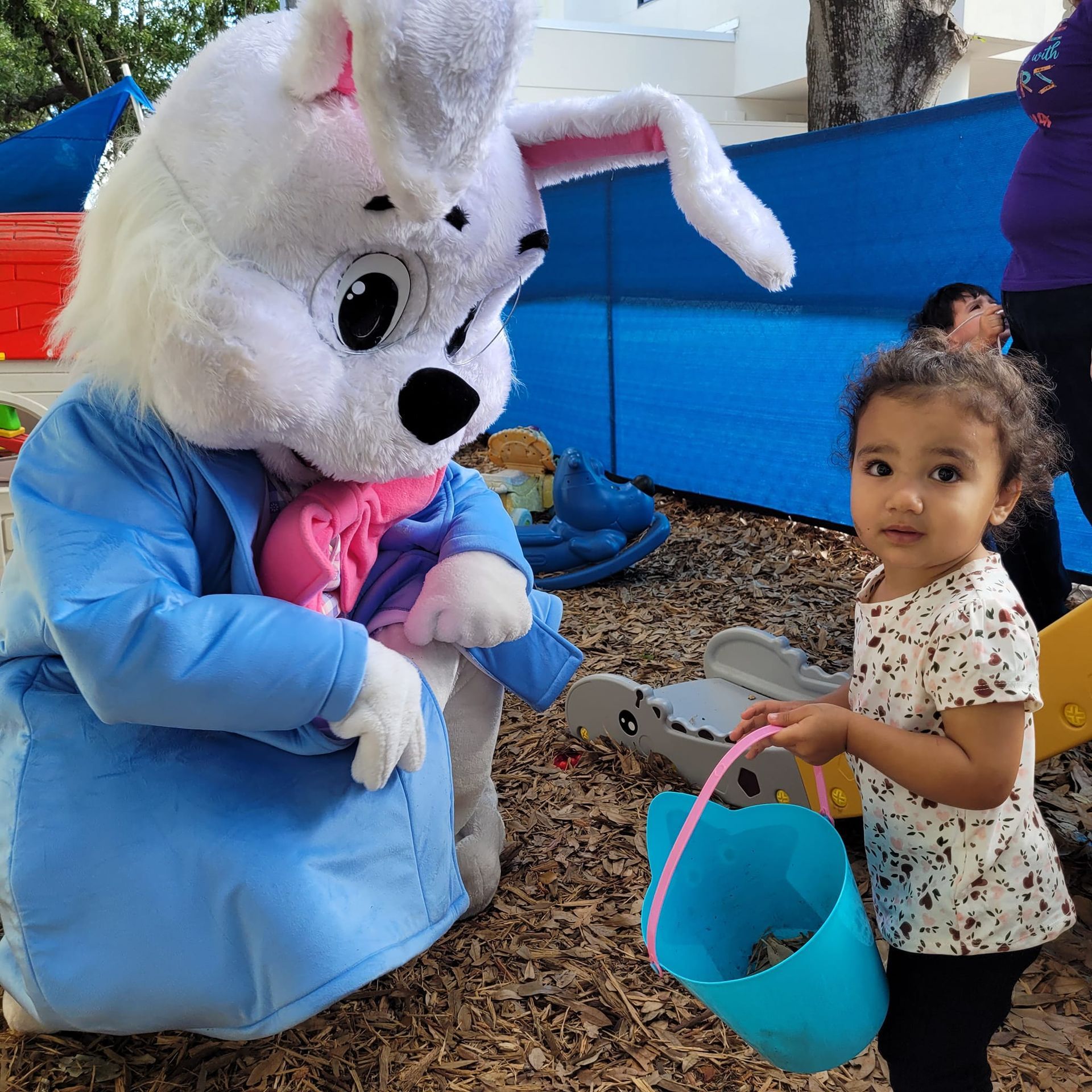 A little girl is holding a blue bucket next to a stuffed bunny