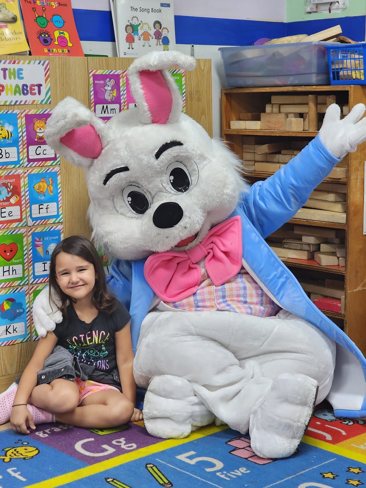 A little girl is sitting next to a bunny mascot in a classroom