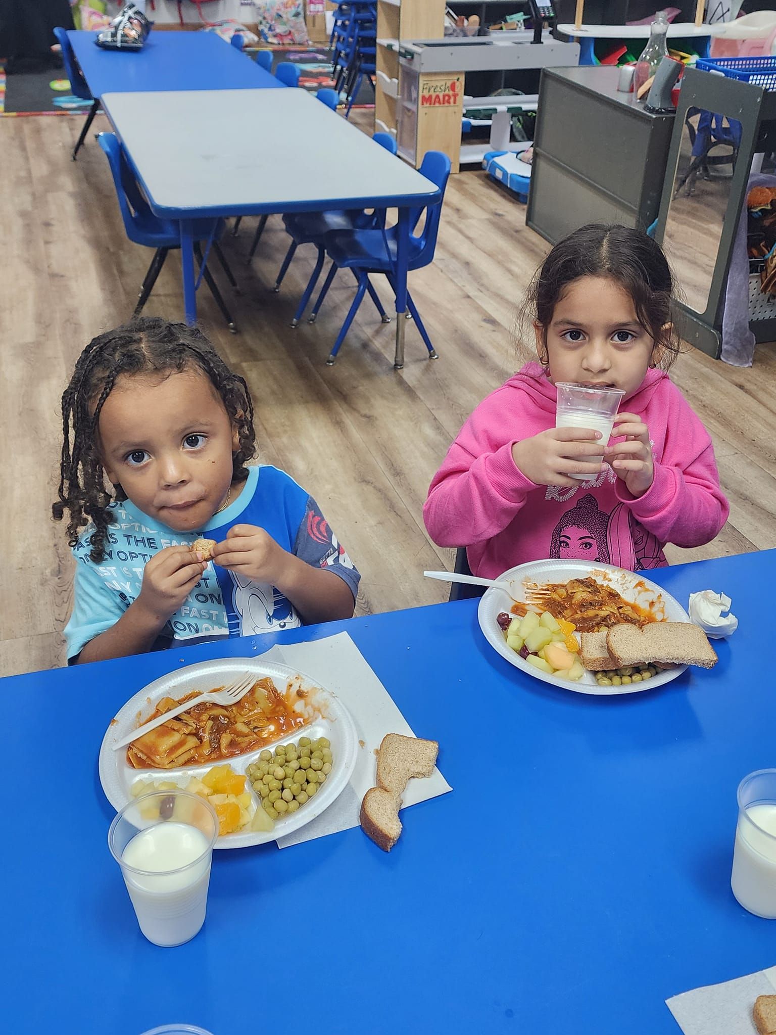 Two little girls are sitting at a table eating food and drinking milk.