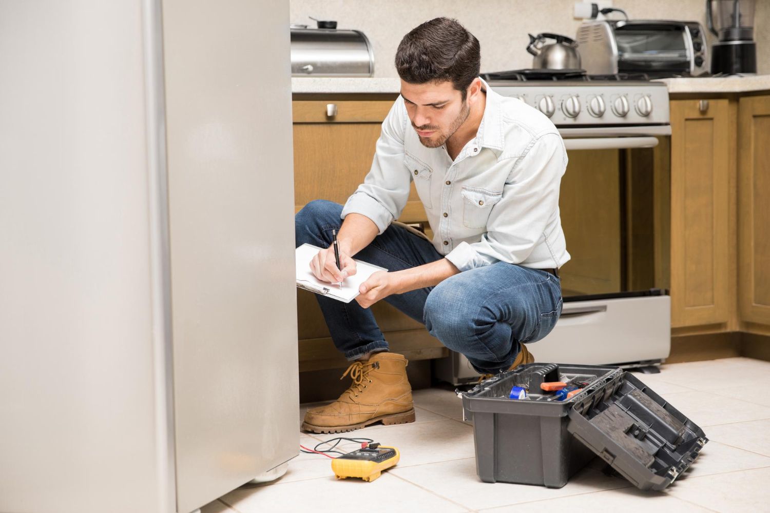 A man is kneeling on the floor in a kitchen repairing a refrigerator.