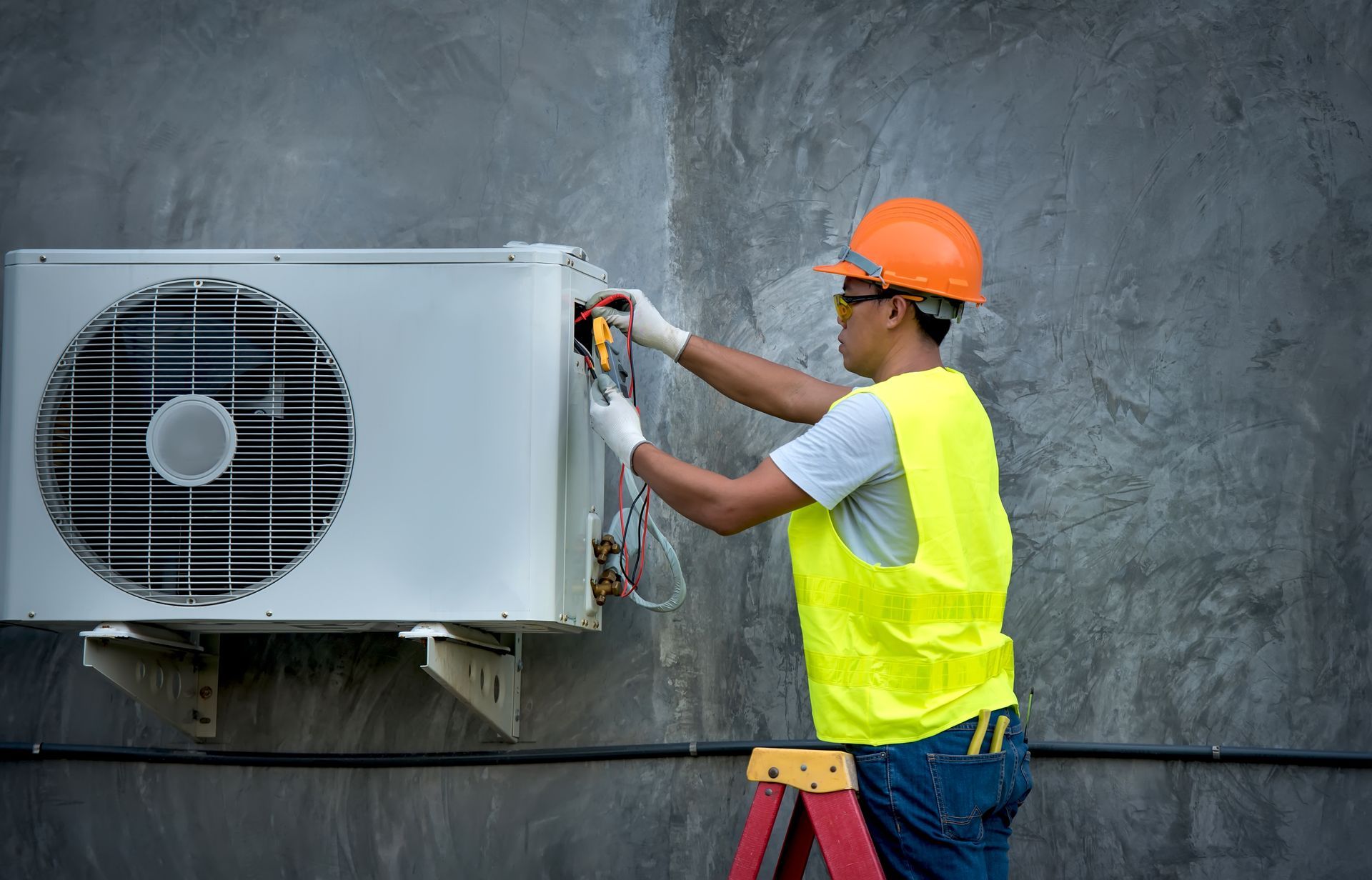 A man is standing on a ladder fixing an air conditioner.